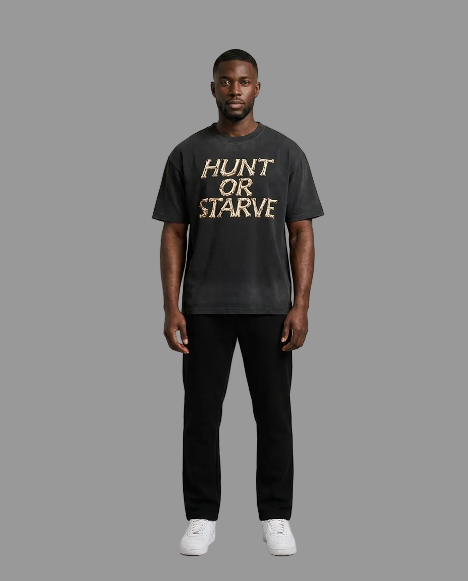 A man modeling a dark charcoal gray graphic t-shirt with 'HUNT OR STARVE' printed in a textured gold font across the chest, paired with black pants and white sneakers, against a neutral beige studio background.