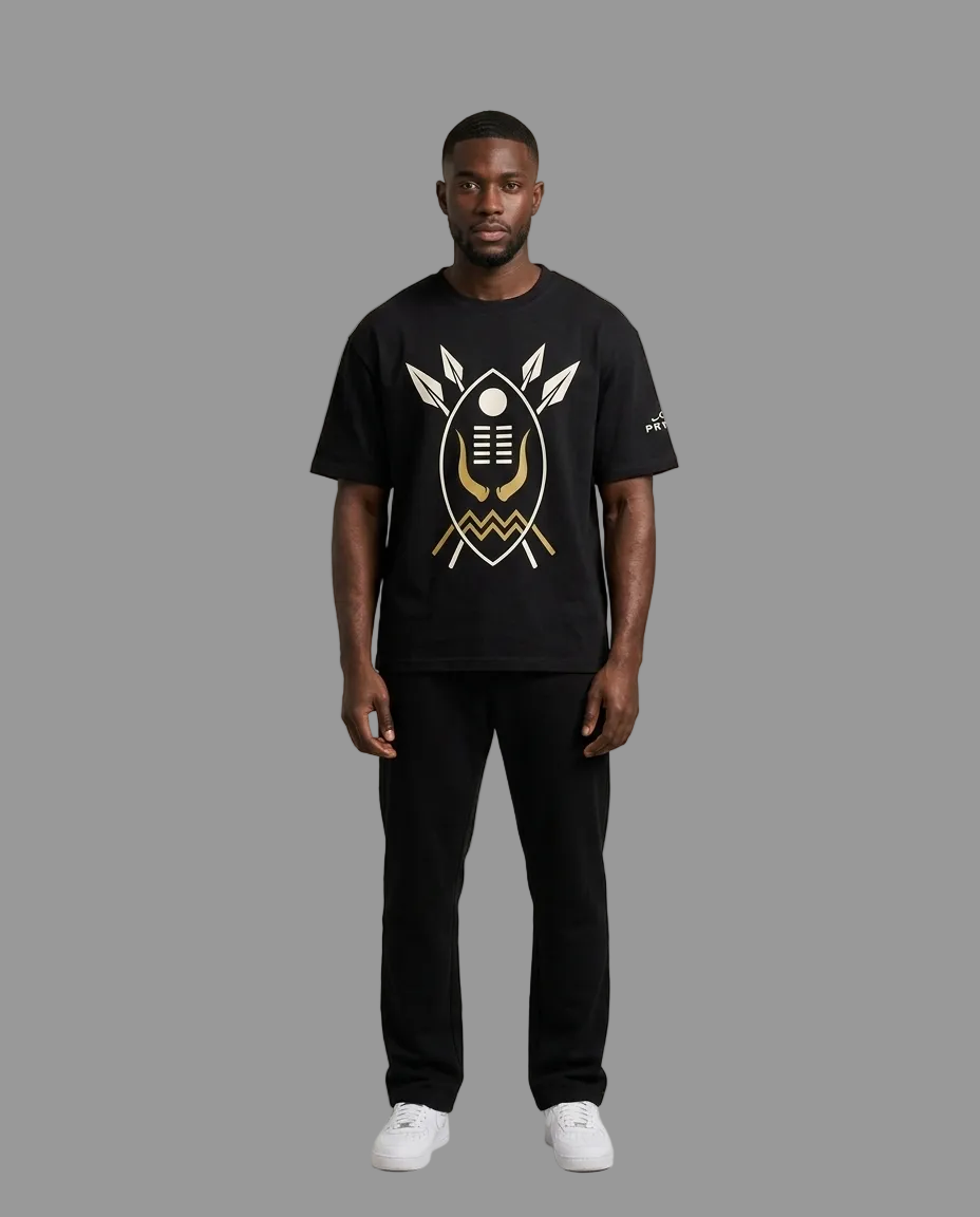 A man modeling a black short-sleeve t-shirt with a large gold and white graphic print on the front, paired with black pants and white sneakers, against a neutral grey studio background.