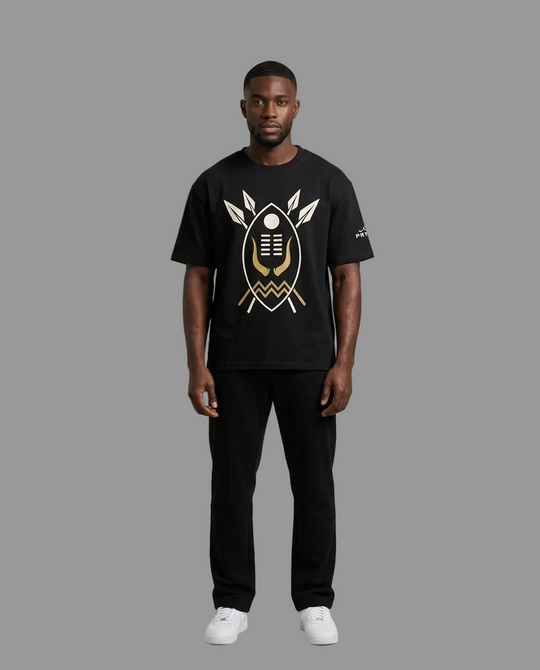 A man modeling a black short-sleeve t-shirt with a large gold and white graphic print on the front, paired with black pants and white sneakers, against a neutral grey studio background.