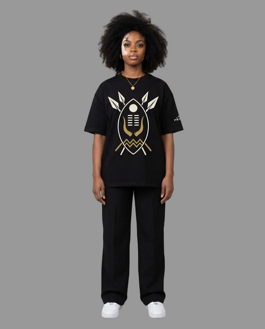 A model stands against a neutral grey studio background wearing a black graphic t-shirt with a gold and white design, black wide-leg pants, and white sneakers.
