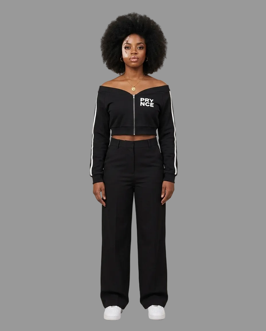 A model with an afro hairstyle stands against a neutral beige studio background wearing a black off-the-shoulder cropped zip-up top with white 'PYNCE' lettering on the chest and white side stripes, paired with black wide-leg pants and white sneakers.