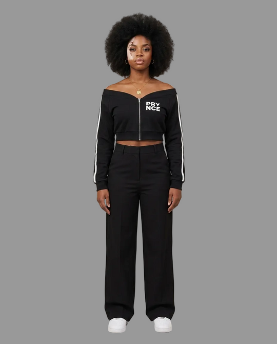 A model with an afro hairstyle stands against a neutral beige studio background wearing a black off-the-shoulder cropped zip-up top with white &