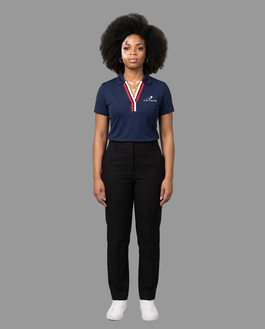 A woman stands against a plain beige studio background wearing a navy polo shirt with a red and white striped collar trim and white logo on the left chest, paired with black trousers and white sneakers.