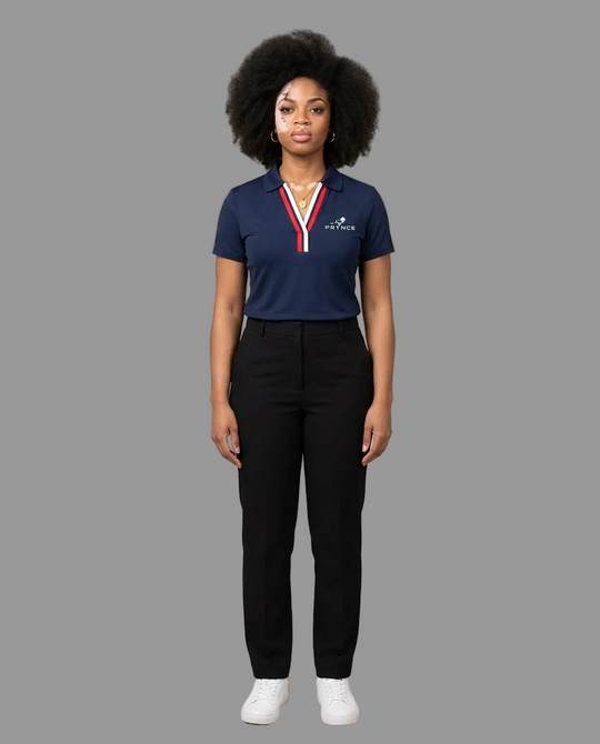 A woman stands against a plain beige studio background wearing a navy polo shirt with a red and white striped collar trim and white logo on the left chest, paired with black trousers and white sneakers.