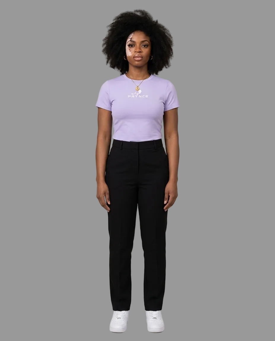A woman stands against a plain beige studio background wearing a light lavender short-sleeve t-shirt with a small white graphic on the chest, black pants, and white sneakers.