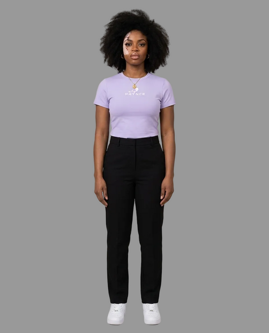 A woman stands against a plain beige studio background wearing a light lavender short-sleeve t-shirt with a small white graphic on the chest, black pants, and white sneakers.