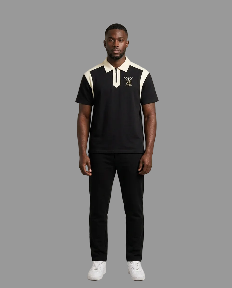 A man modeling a black short-sleeve polo shirt with cream-colored shoulder panels and collar, featuring a small embroidered crest on the left chest, paired with black trousers and white sneakers against a neutral studio background.