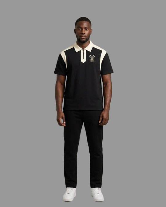 A man modeling a black short-sleeve polo shirt with cream-colored shoulder panels and collar, featuring a small embroidered crest on the left chest, paired with black trousers and white sneakers against a neutral studio background.