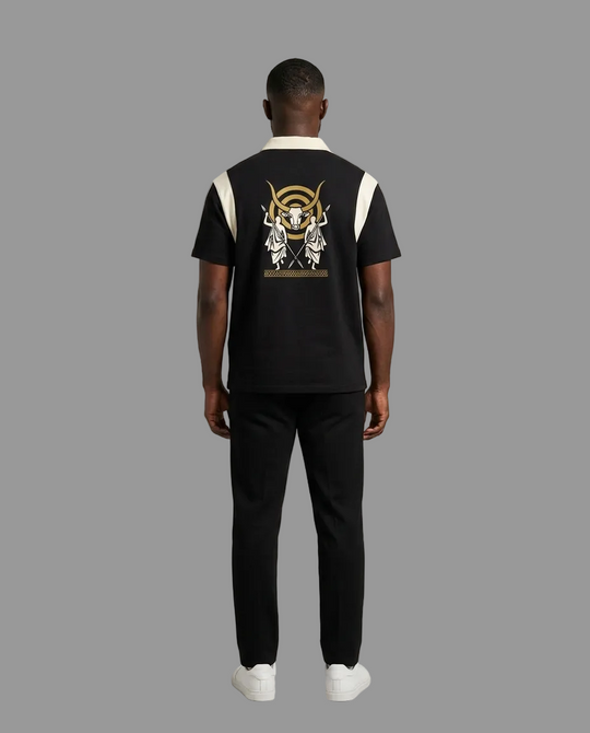 A man viewed from the back wearing a black short-sleeve polo shirt with white shoulder panels and a large gold and white graphic print on the back, paired with black pants and white sneakers, against a neutral studio background.