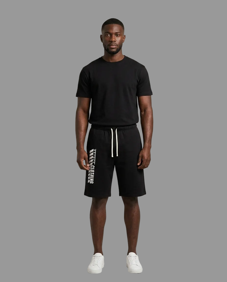 A male model stands against a light beige studio background wearing a black short-sleeve t-shirt, black athletic shorts with white drawstrings and vertical white text, and white sneakers.