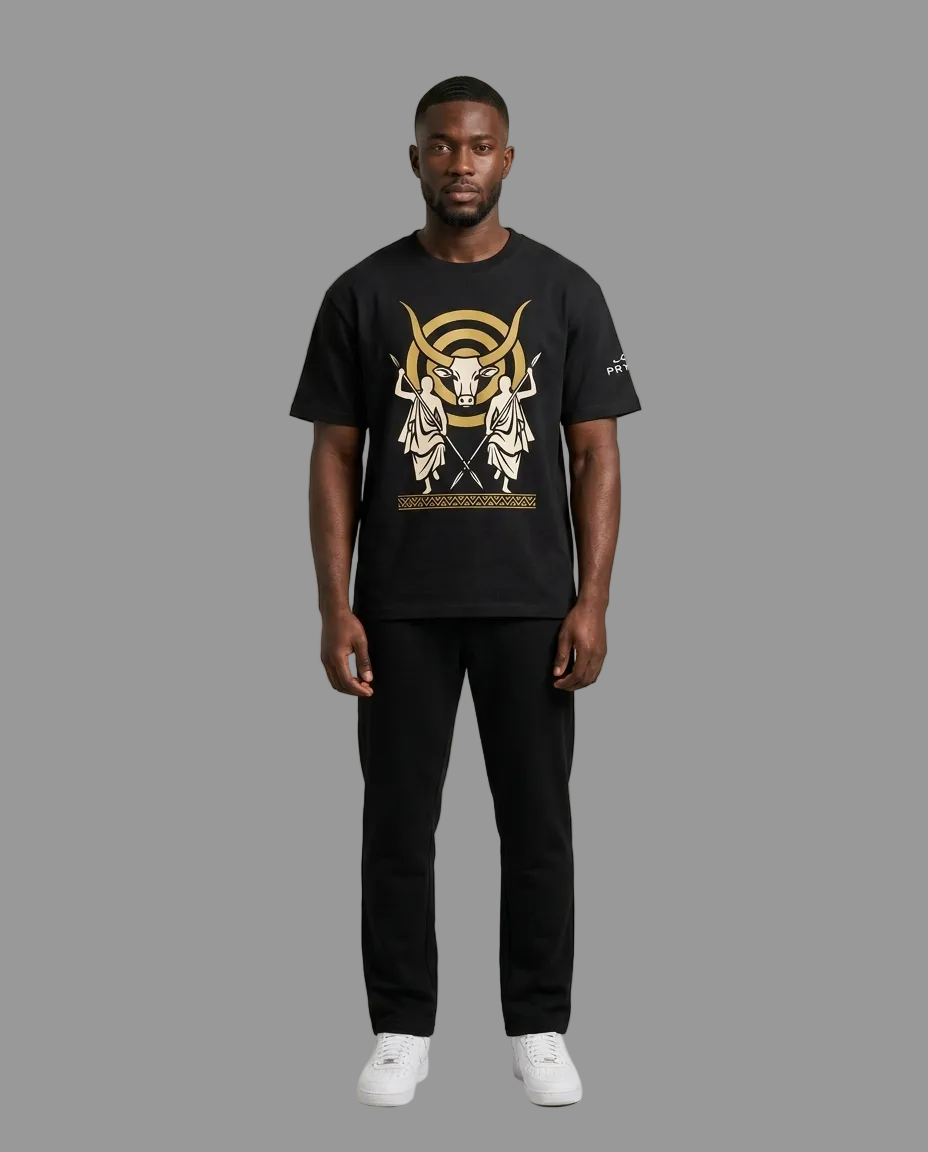 A man modeling a black short-sleeve t-shirt with a large gold and white graphic print on the front, paired with black pants and white sneakers, against a neutral light gray background.