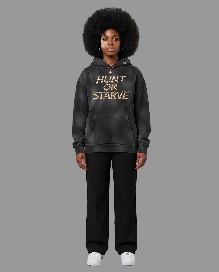 A model wearing a washed black pullover hoodie with 'HUNT OR STARVE' printed in gold across the chest, paired with black pants and white sneakers, set against a plain beige studio background.