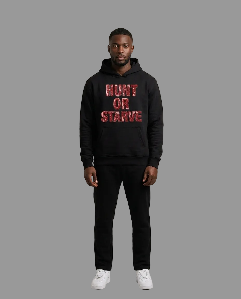 A man modeling a black pullover hoodie with 'HUNT OR STARVE' printed in red distressed letters across the chest, paired with black pants and white sneakers, against a neutral studio background.