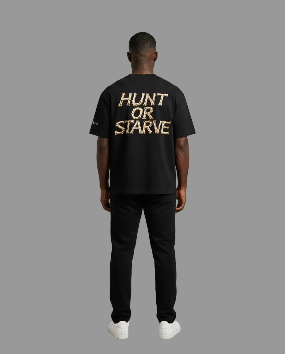 A man viewed from behind wearing a black t-shirt with 'HUNT OR STARVE' printed in gold across the back, paired with black pants and white sneakers, against a neutral studio background.