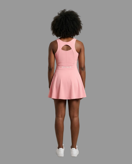 A model viewed from behind wearing a pink sleeveless athletic dress with a keyhole cutout at the back and a flared skirt, paired with white sneakers, against a solid light grey background.