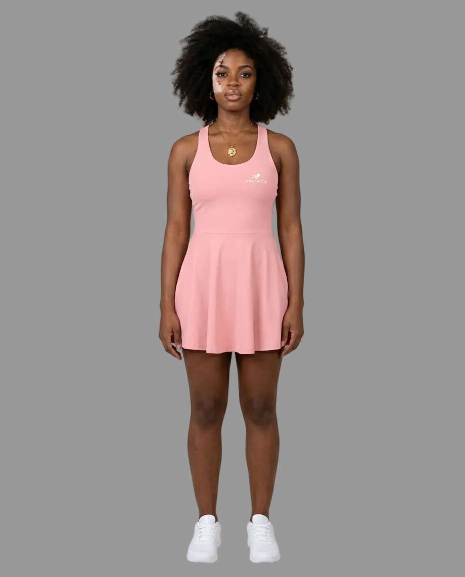 A woman modeling a light pink sleeveless athletic dress with a small white logo on the left chest, paired with white sneakers, against a solid light grey studio background.