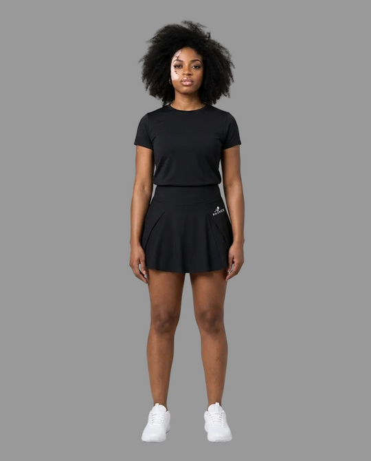 A woman modeling a black short-sleeve athletic t-shirt and matching black tennis skirt with white sneakers, standing against a light gray studio background.