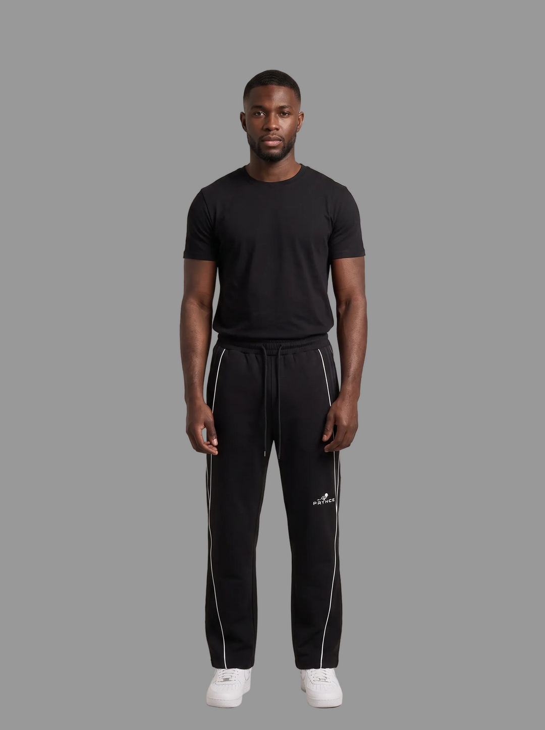 A man modeling a black short-sleeve t-shirt and black sweatpants with white side stripes and a white Adidas logo, standing against a solid grey studio background.