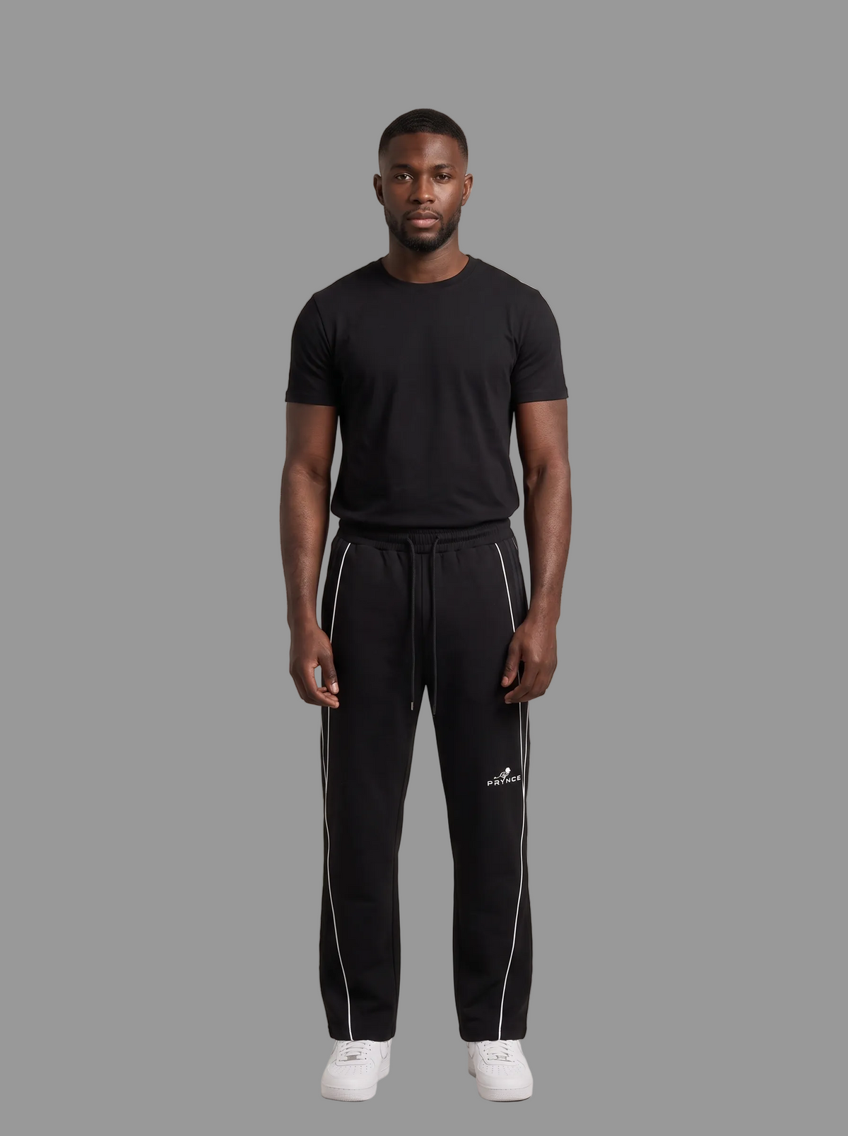 A man modeling a black short-sleeve t-shirt and black sweatpants with white side stripes and a white Adidas logo, standing against a solid grey studio background.